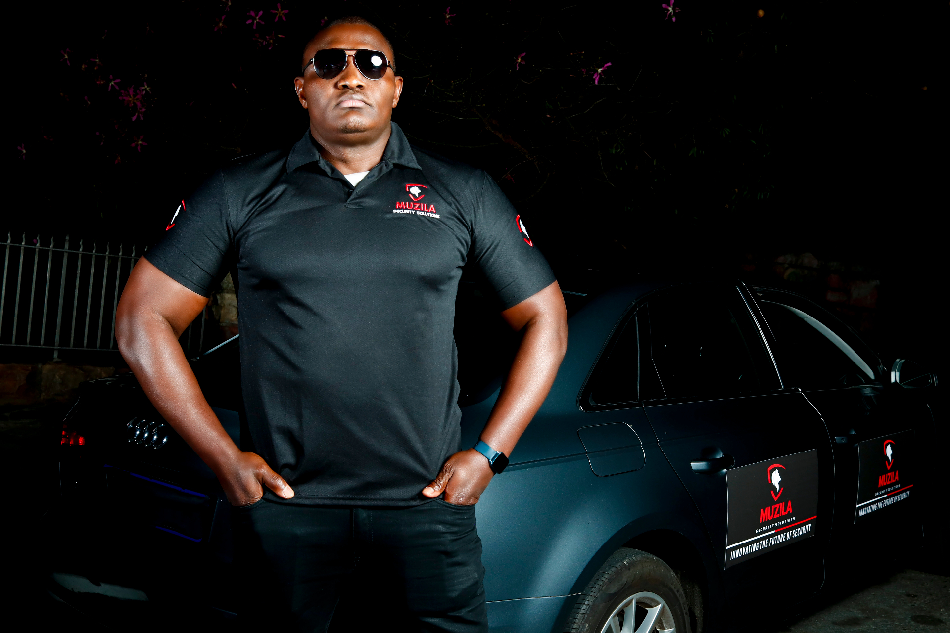 A Muzila close-protection officer in tactical kit standing watch at night, framed by low ambient light.